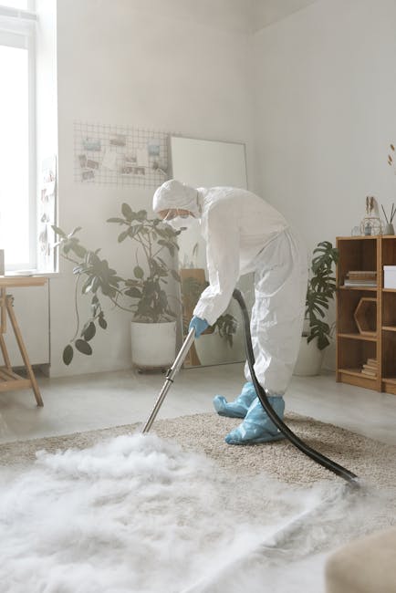 A professional cleaning technician in a white protective suit, face mask, and gloves, using a vacuum or steaming device to deep clean and sanitise a beige carpet in a bright, modern living room. The room features white walls, a wooden side table with decorative items, large green potted plants, and natural light coming through a window, highlighting the cleanliness and shine of the carpet. This scene exemplifies surface cleaning and deep cleaning services offered by Ilford Cleaner, demonstrating thorough domestic cleaning processes in a residential setting.