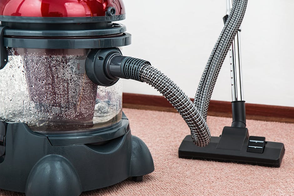 Close-up of a carpet cleaning vacuum in action on a pinkish-beige carpet, with a transparent dust container filled with debris and foam visible, indicating recent cleaning. The black and grey vacuum hose is connected to a cleaning head positioned on the floor. The background shows a plain white wall and a wooden skirting board, with the scene illuminated by natural or artificial light, highlighting the cleanliness and shine of the carpet surface. The image emphasizes surface cleaning and deep cleaning processes, exemplifying professional domestic cleaning services provided by Ilford Cleaner, which specializes in hygiene and maintenance.