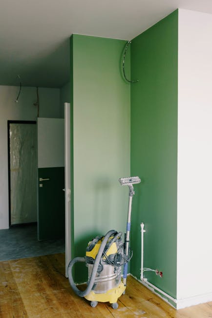 A room in the process of renovation cleaning featuring a wooden floor and green walls, with a partially visible doorway leading to another space with a gray tiled floor and white walls. The room conta