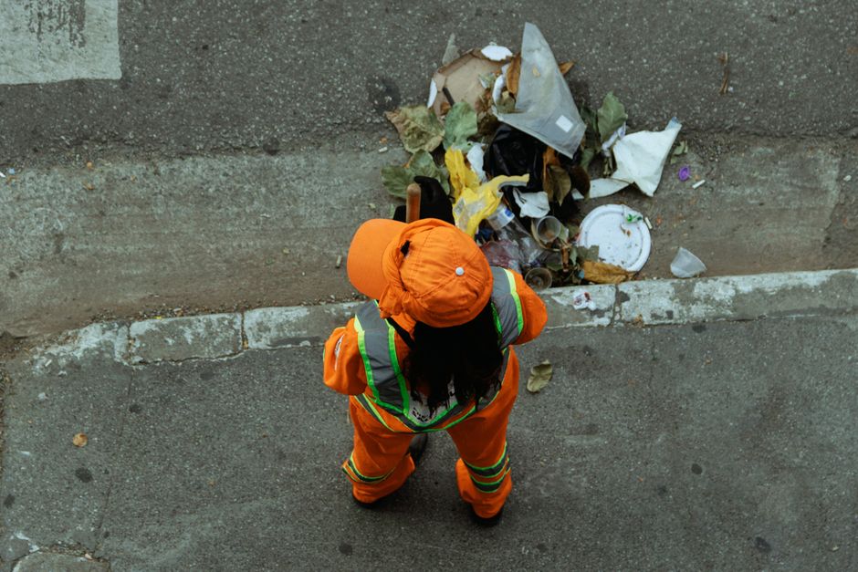 A waste management worker dressed in an orange high-visibility uniform and matching hat stands on a paved street, facing a large pile of mixed rubbish including paper, plastic, cardboard, and leaves,