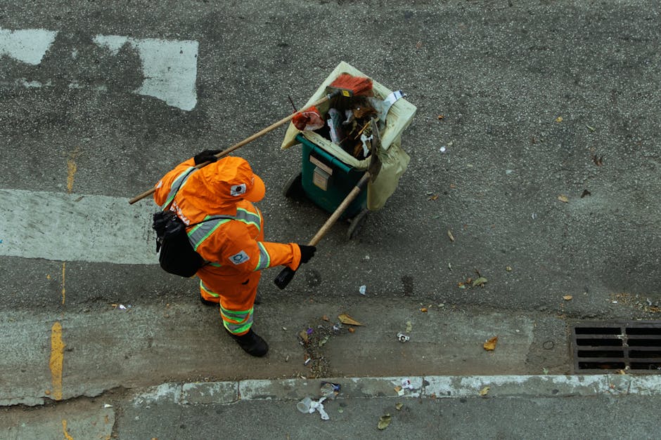 A waste management worker dressed in an orange high-visibility uniform and matching hat stands on a paved street, facing a large pile of mixed rubbish including paper, plastic, cardboard, and leaves,