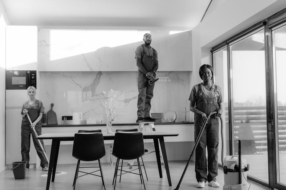 Three professional cleaners are working in a modern kitchen for surface cleaning and deep cleaning. One woman on the left is holding a mop, standing near a black bucket, while a man in the center is standing on a step ladder, wiping a white marble backsplash on the wall. Another woman on the right is mopping the wooden floor, with a vacuum cleaner nearby. The kitchen features a sleek white marble countertop, a built-in microwave, and large sliding glass doors allowing natural light to illuminate the space. The lighting highlights the spotless and shiny surfaces, indicating thorough cleaning and sanitisation by Ilford Cleaner, specializing in domestic cleaning services near Ilford station.