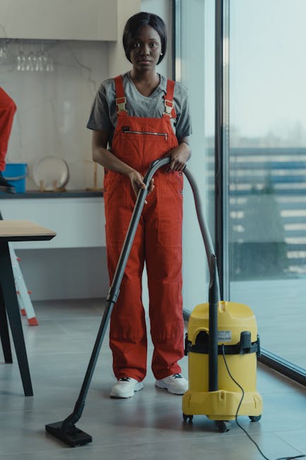 A young woman with dark hair, wearing a grey t-shirt and red overalls, is operating a yellow commercial vacuum cleaner with a black hose in a modern, well-lit kitchen. The kitchen features a light wood floor, white cabinetry, and a grey countertop with minimal accessories. Large floor-to-ceiling glass windows and doors reveal a balcony with a metal railing outdoors. The surface area appears clean, with the vacuum cleaner in use for deep cleaning or surface hygienic maintenance, suggesting professional domestic or commercial cleaning services by Ilford Cleaner. The overall scene emphasizes thorough cleaning, hygiene, and surface sanitisation efforts in a contemporary residential setting.