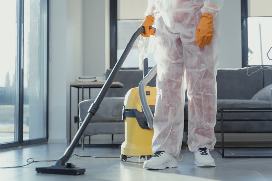 Three professional cleaners are working in a modern kitchen for surface cleaning and deep cleaning. One woman on the left is holding a mop, standing near a black bucket, while a man in the center is standing on a step ladder, wiping a white marble backsplash on the wall. Another woman on the right is mopping the wooden floor, with a vacuum cleaner nearby. The kitchen features a sleek white marble countertop, a built-in microwave, and large sliding glass doors allowing natural light to illuminate the space. The lighting highlights the spotless and shiny surfaces, indicating thorough cleaning and sanitisation by Ilford Cleaner, specializing in domestic cleaning services near Ilford station.