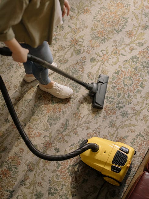 A yellow vacuum cleaner with a black hose on a patterned area rug in a living room corner, next to a wooden console table. The console holds a white bust sculpture, a remote control, a chessboard, and