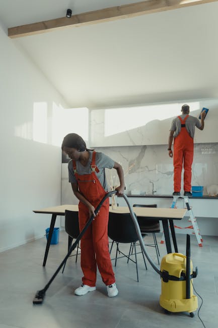 A monochrome image shows three professional cleaners in a modern kitchen, engaged in surface cleaning and maintenance. One cleaner stands on a step ladder, wiping the white marble backsplash with a cl